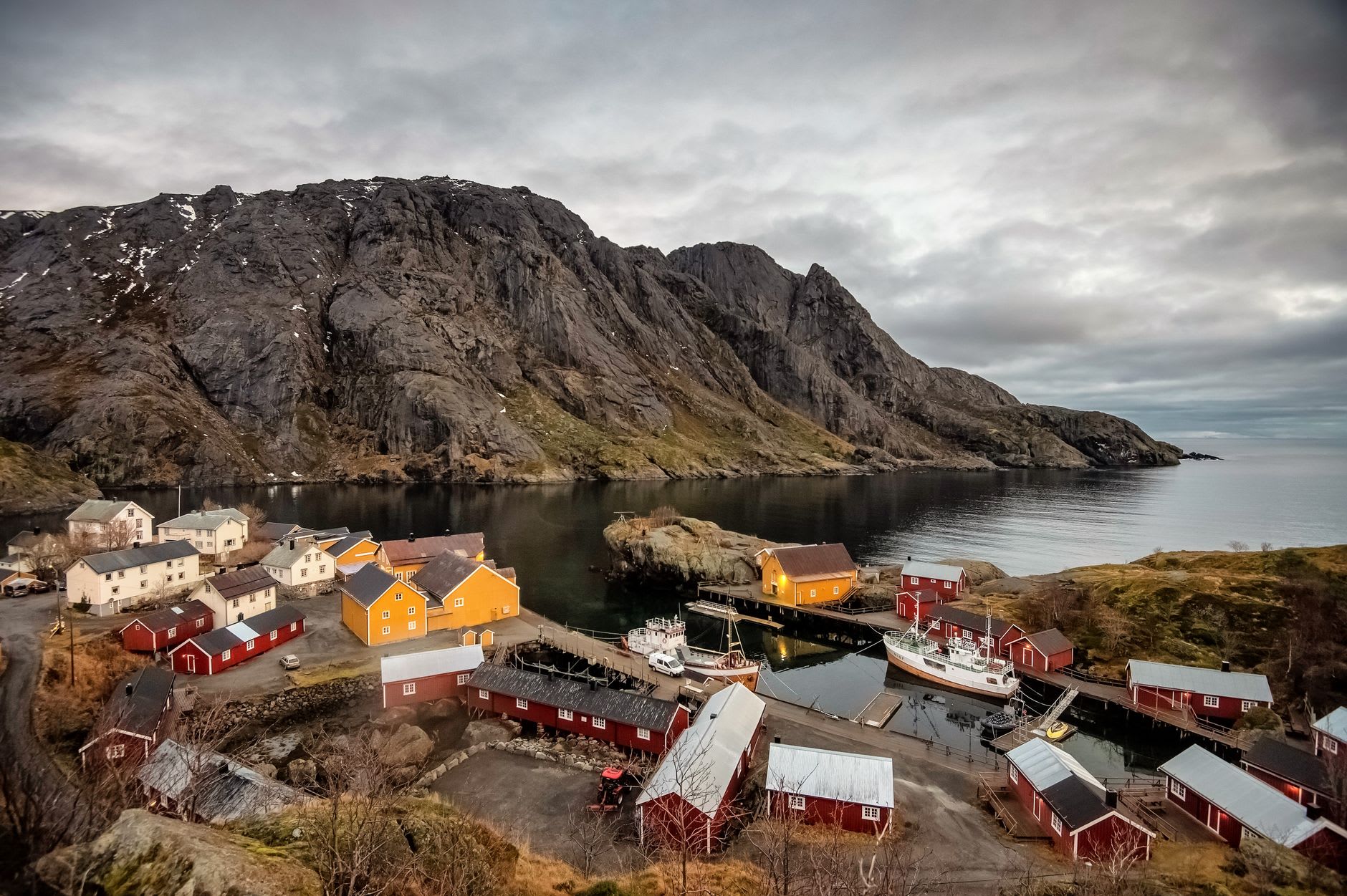 Nusfjord boathouses reflected in the still fjord at low tide on a grey morning, rorbu walls weathered to deep red