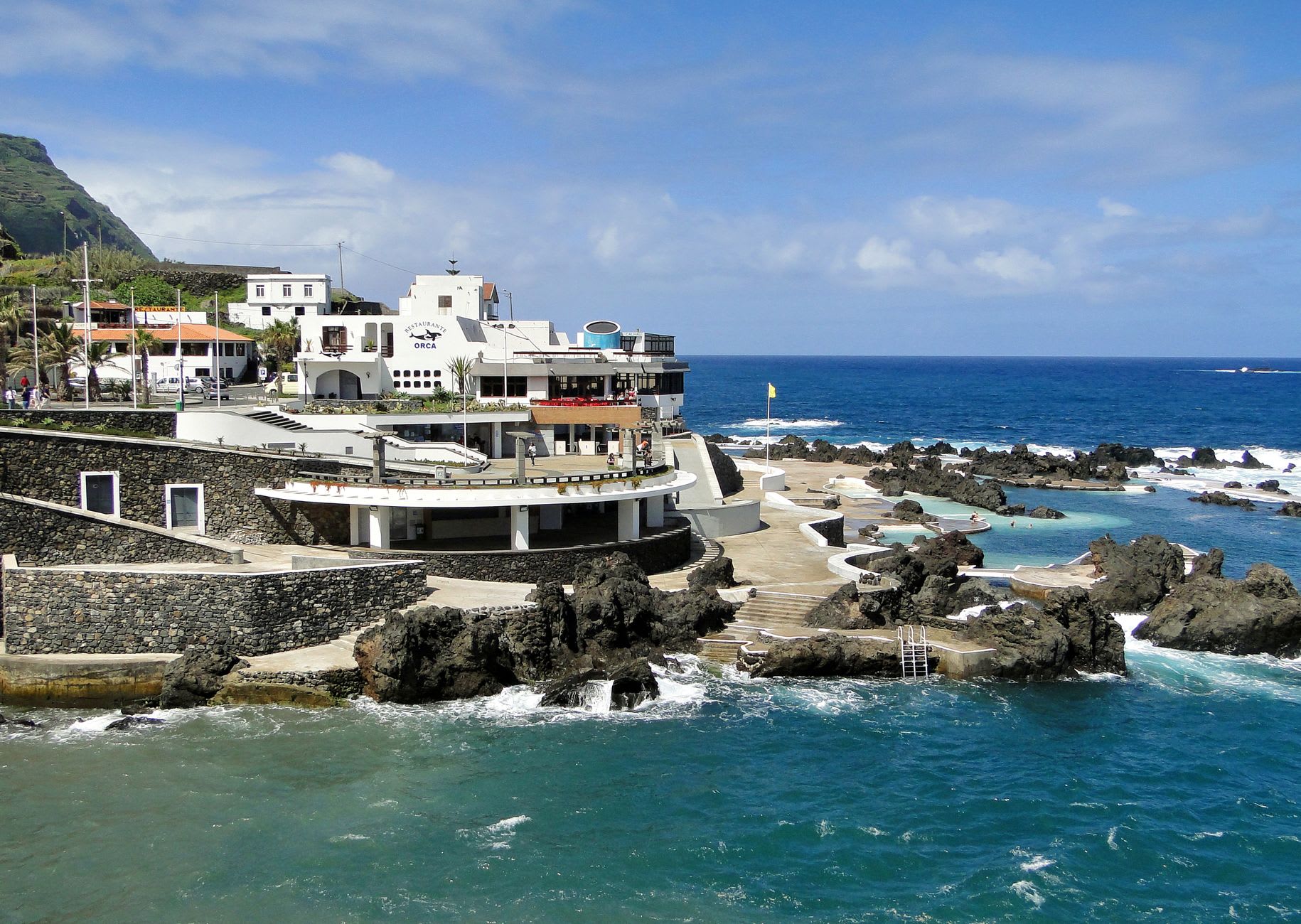 Swimmers in the emerald-green volcanic lava pools at Porto Moniz, Atlantic waves crashing against the outer sea wall