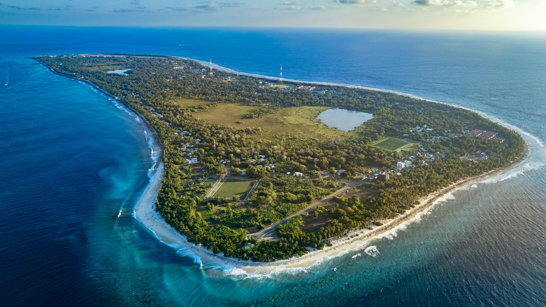 The reef edge of Kwajalein Atoll from a low-altitude approach, the lagoon's color gradient shifting from cobalt blue to translucent jade over the coral shelf