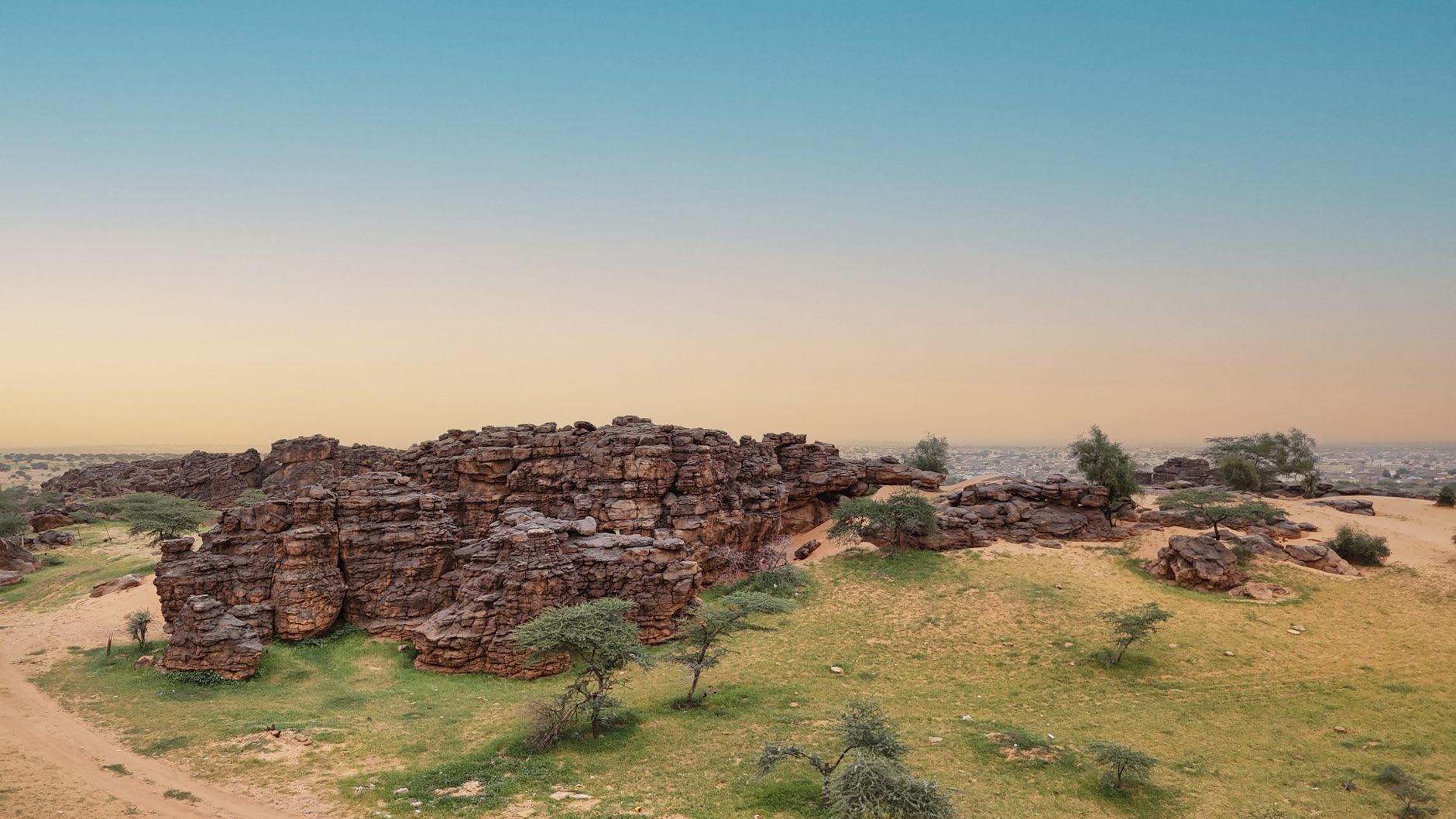 Travellers silhouetted on top of a heaped iron-ore wagon as the train crosses empty Saharan desert at dusk near Zouérat, the line of wagons curving away to the horizon