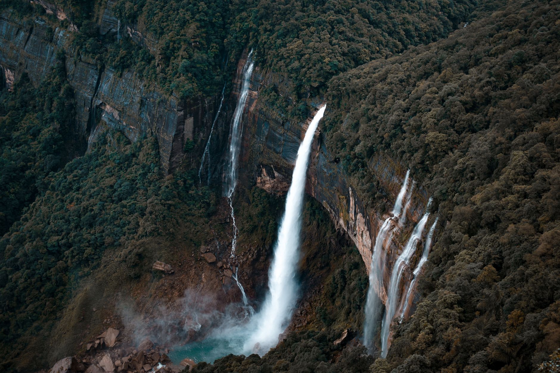 The small emerald pool at the base of Nohkalikai Falls seen through telephoto lens, mist rising from where the water strikes the surface