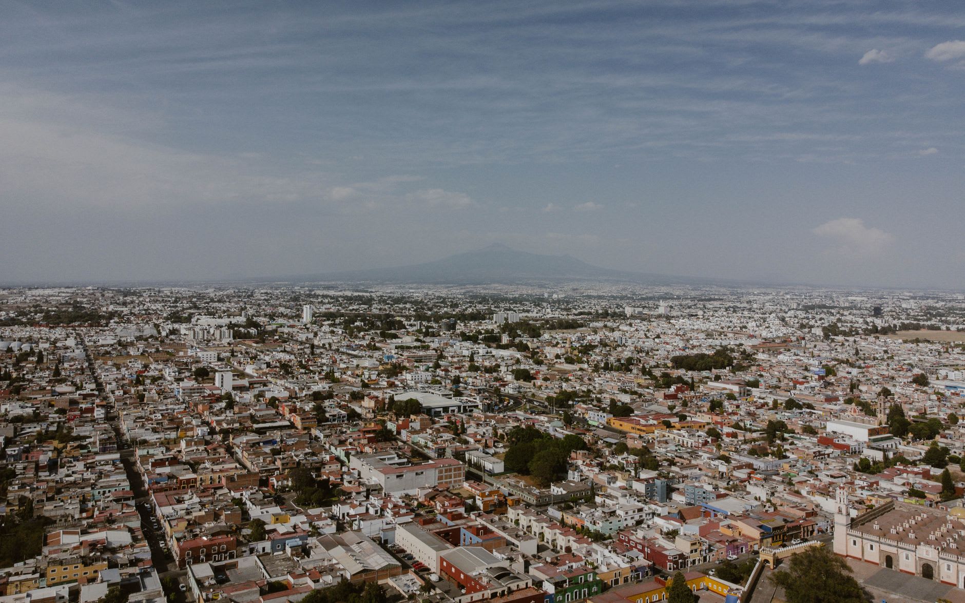 The Volcán de Fuego visible above the red-tiled rooftops of Colima city at dawn, the volcanic cone perfectly formed against the pale early sky, a thin white plume of vapor at the summit