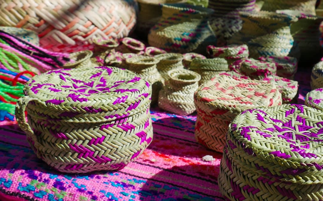 A Rarámuri woman weaving a pine needle basket at a craft market in Creel, Chihuahua, her traditional brightly colored dress visible, the finished baskets displayed beside her, the pine forest visible behind the market stalls