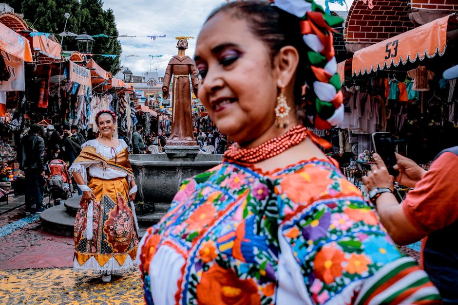 The Sunday tianguis market in Cuetzalan's plaza, Nahua women in traditional white quexquémitl capes selling vegetables and flowers, the colonial church portal visible behind the colorful market stalls