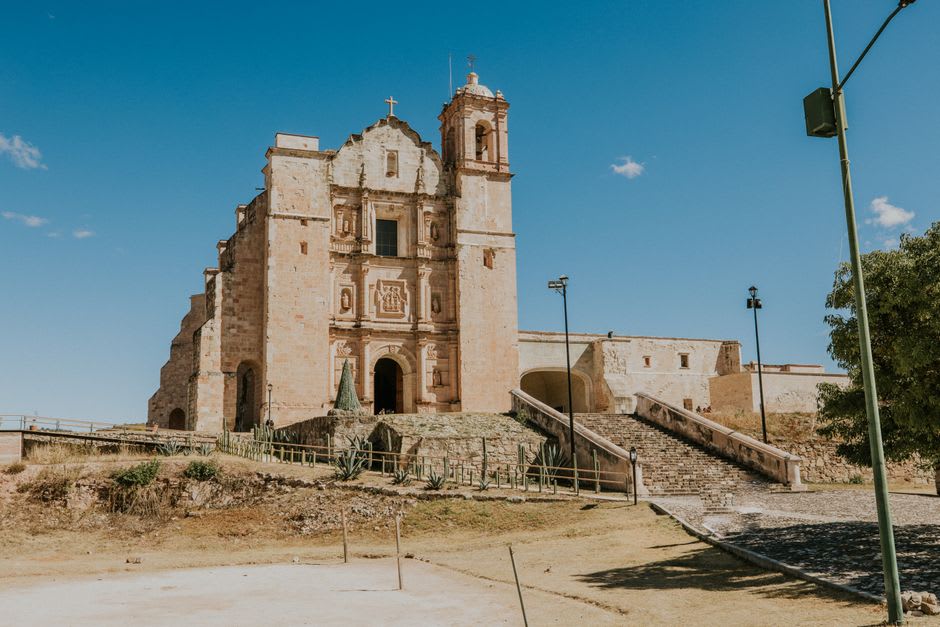 A quiet street in Cuilapan de Guerrero with the stone convent wall visible at the end, a few market stalls along the sidewalk