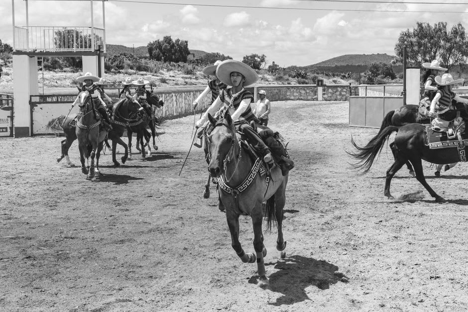 Riders preparing at the chute at the Jesús María jaripeo, families visible in the stands behind