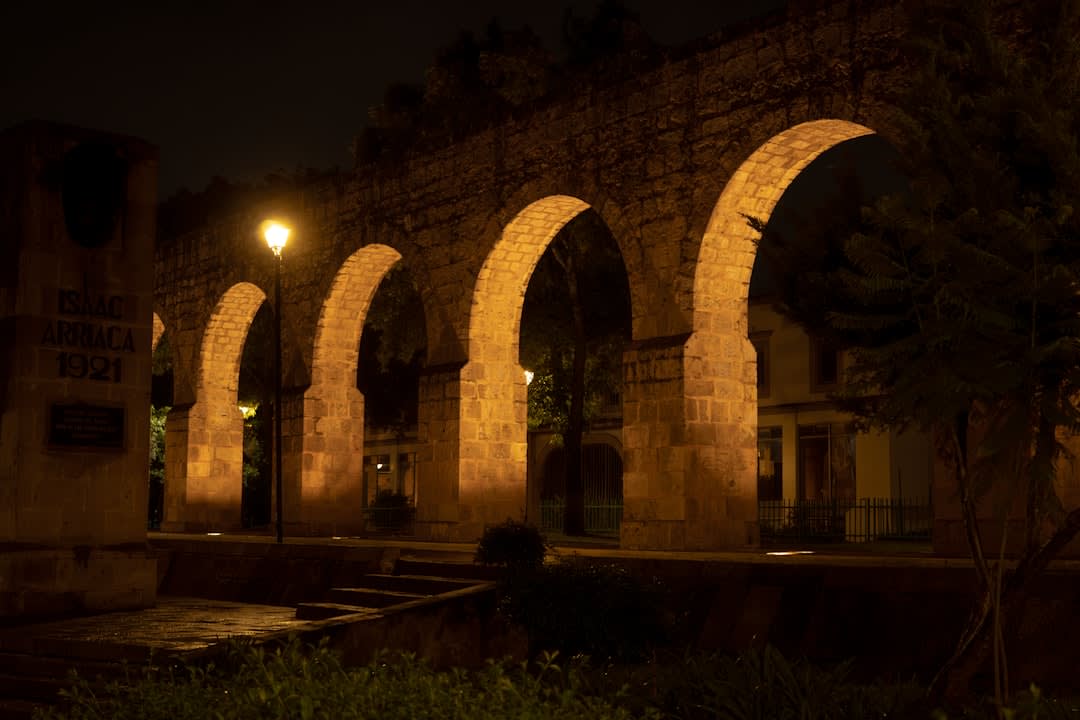 The illuminated pink cantera stone twin towers of Morelia's cathedral at dusk, arched portales and colonial buildings surrounding the Plaza de Armas