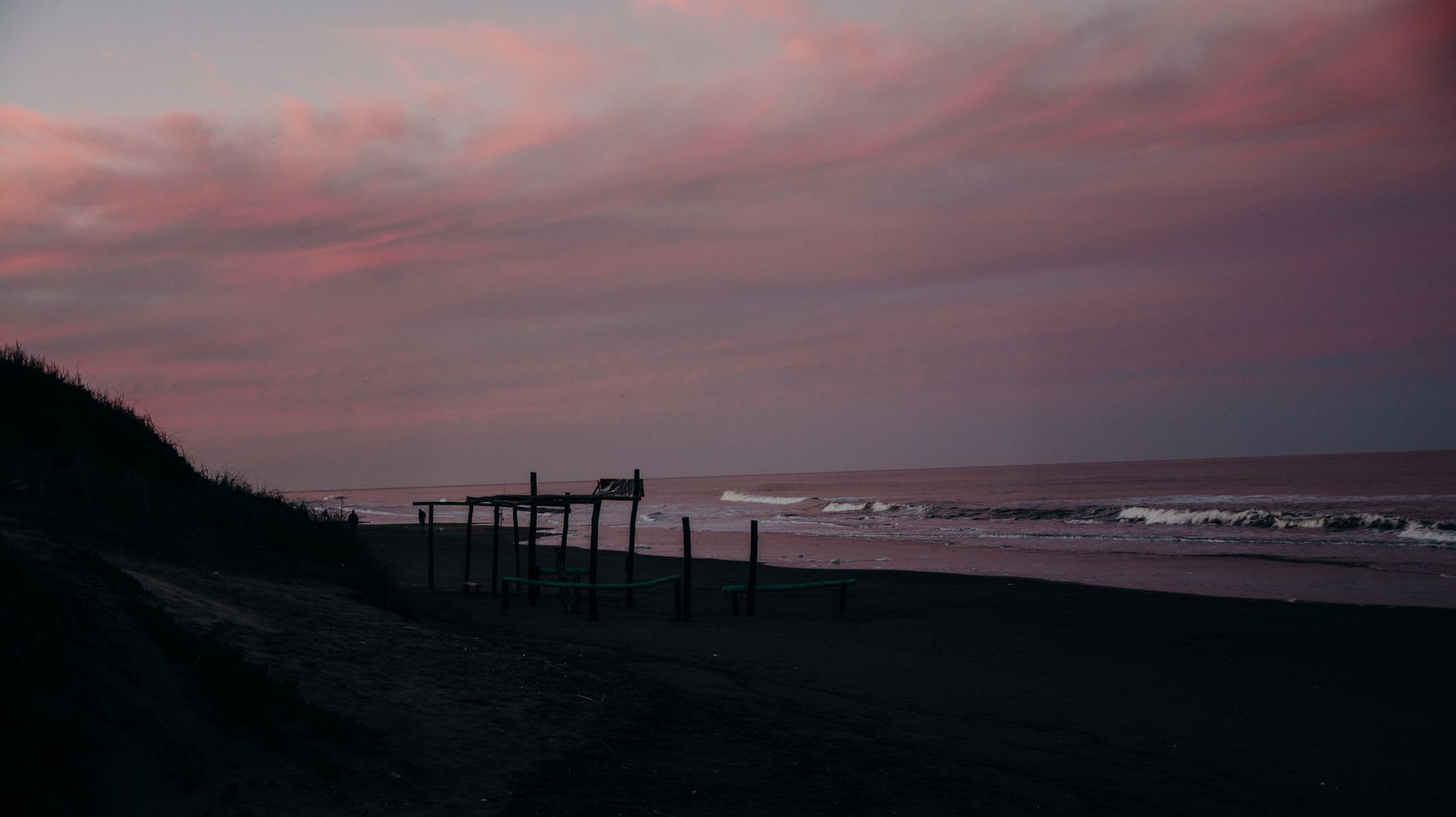 The Pacific beach of Pie de la Cuesta in late afternoon, heavy surf breaking close to a row of palapa restaurants, families at plastic tables in the sand, palm trees bending in the on-shore wind