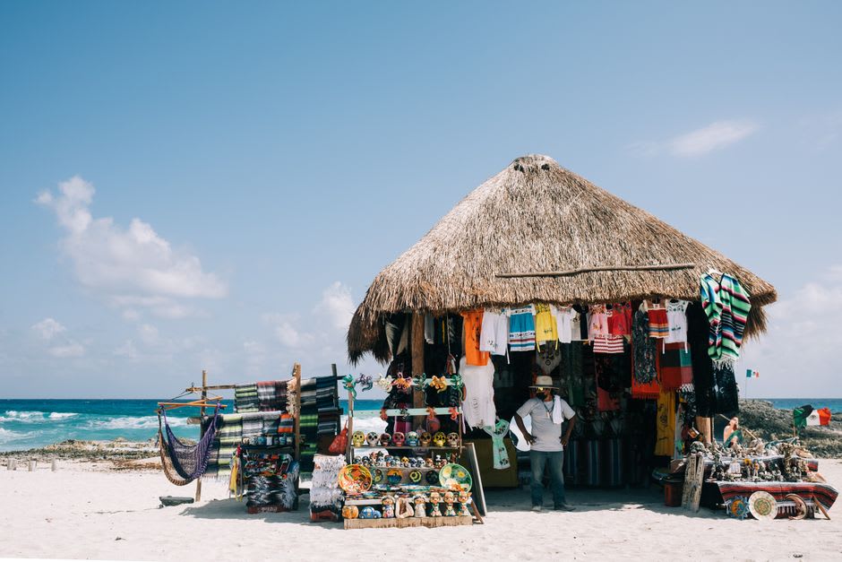 The Puerto Morelos artisan market under a thatched palapa, local craftspeople at their stalls with the main street behind and the sea visible at the end