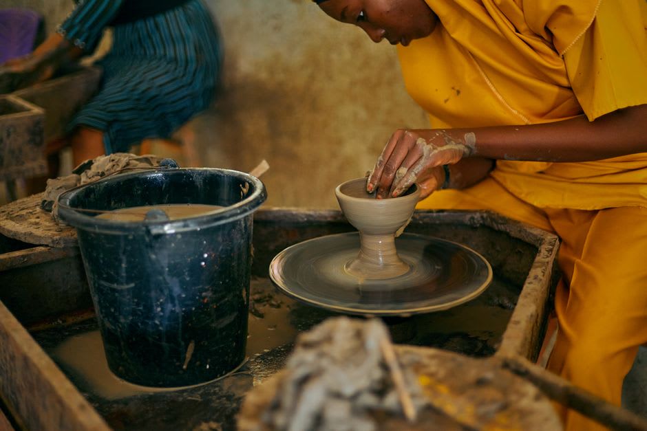 A potter at the Atzompa cooperative working clay on a wheel, green-glazed pieces drying on boards behind her, the workshop space open to the morning light