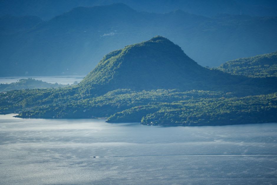 Late afternoon light on the surface of Santa María del Oro lake, the water turned grey-green under clouds, the pine-covered caldera walls reflected in the still surface