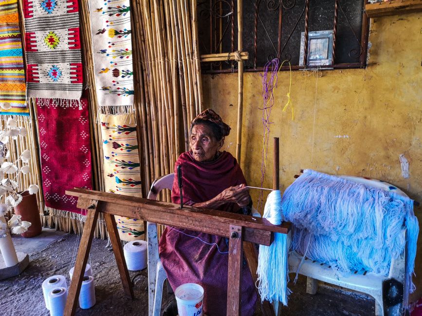 A completed Teotitlán del Valle rug with traditional Zapotec geometric patterns in cochineal red, indigo blue, and marigold yellow, hung on a white wall in the workshop, the weaver's loom visible behind