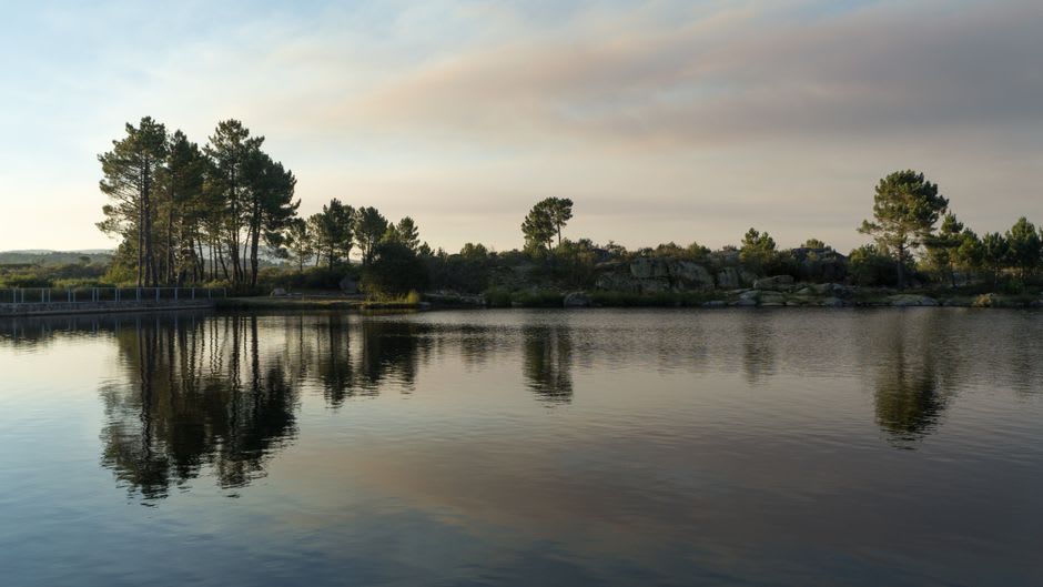 Fishermen in a small boat on the still grey water of the Presa Villa Victoria reservoir, pine forest and farmland on the far shore under a lifting highland mist
