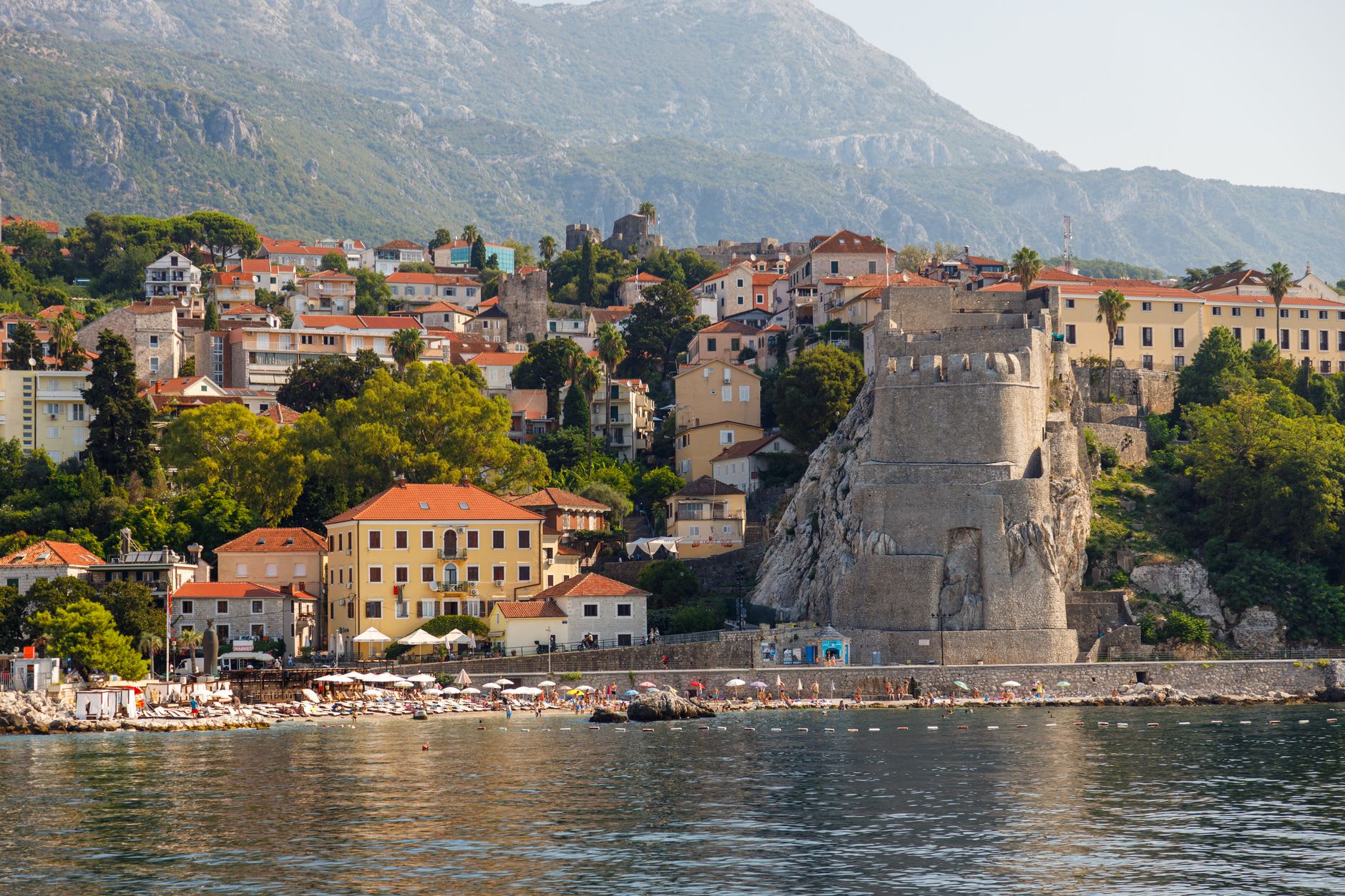 The Šetalište waterfront promenade in late afternoon, palm trees lining the path, the Herceg Novi fortress visible on the hill above