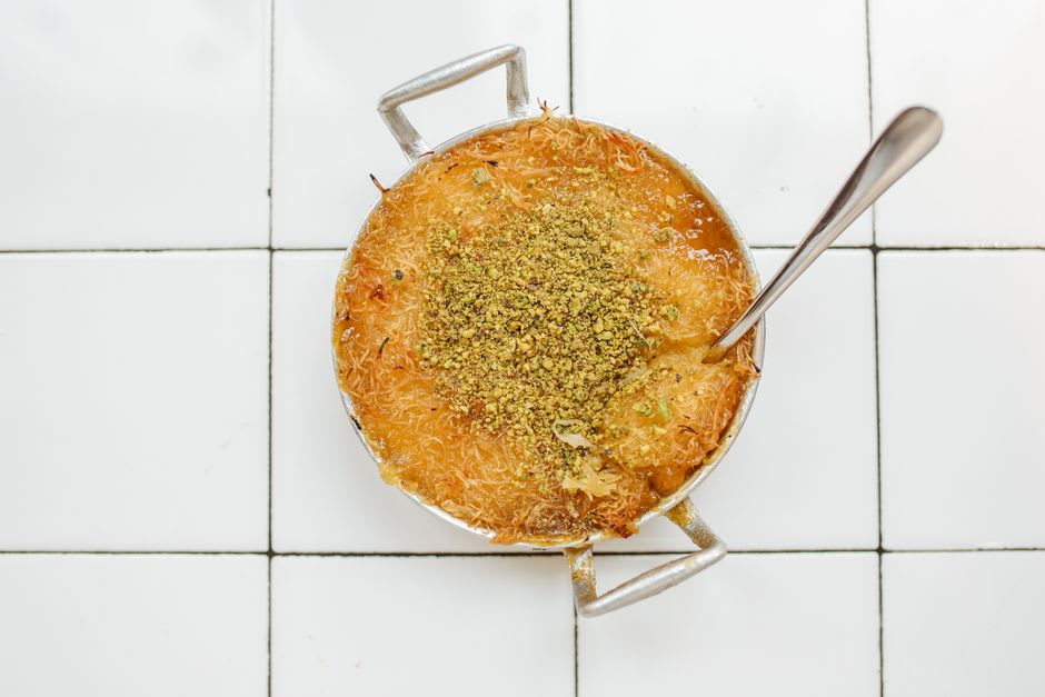 A stall selling fresh knafeh pastry in the old town market of Al-Karak, steam rising from the pan beside the castle walls in the background