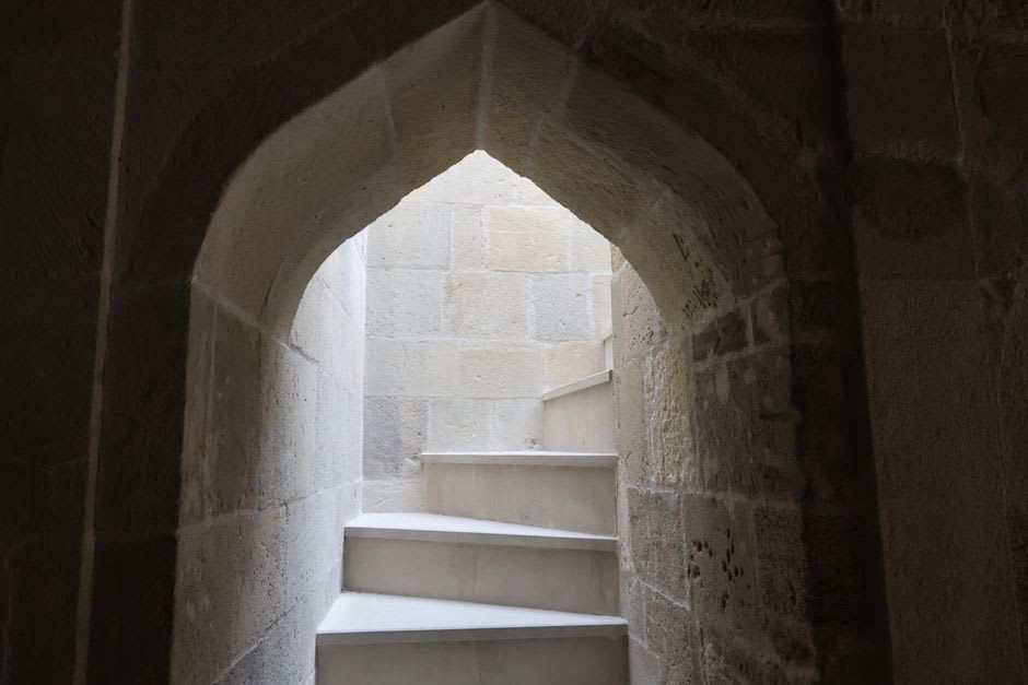 Un guía local descendiendo por la oscura escalera de caracol tallada en la roca bajo el castillo de Shobak, la luz de una antorcha iluminando los antiguos escalones de piedra