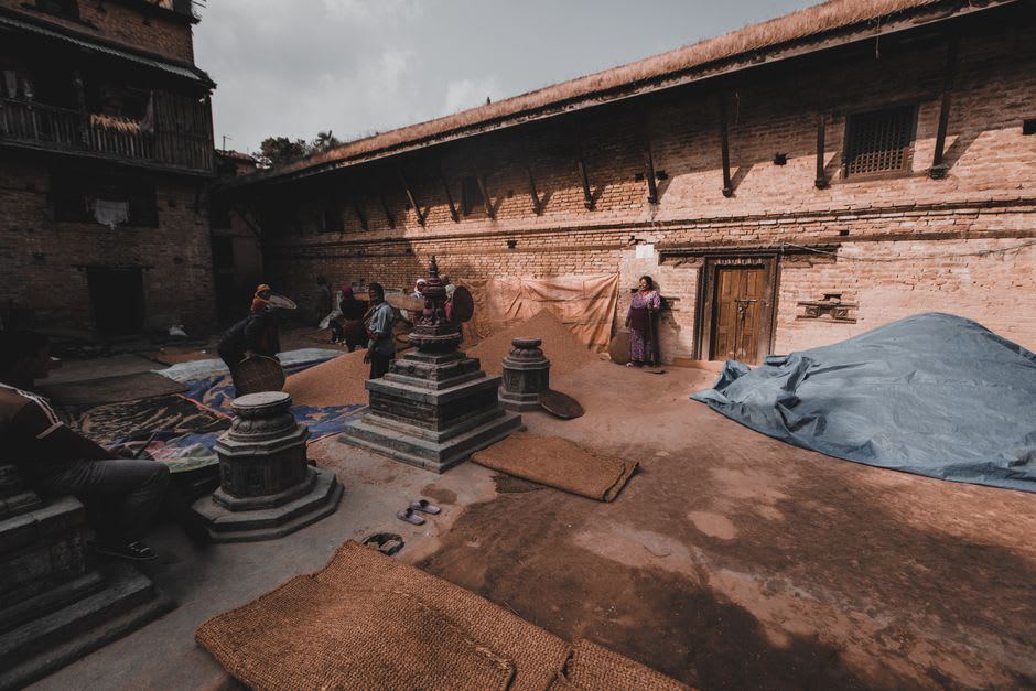 Elderly woman drying grain on the stone plinth of a small temple courtyard in Panauti's old town