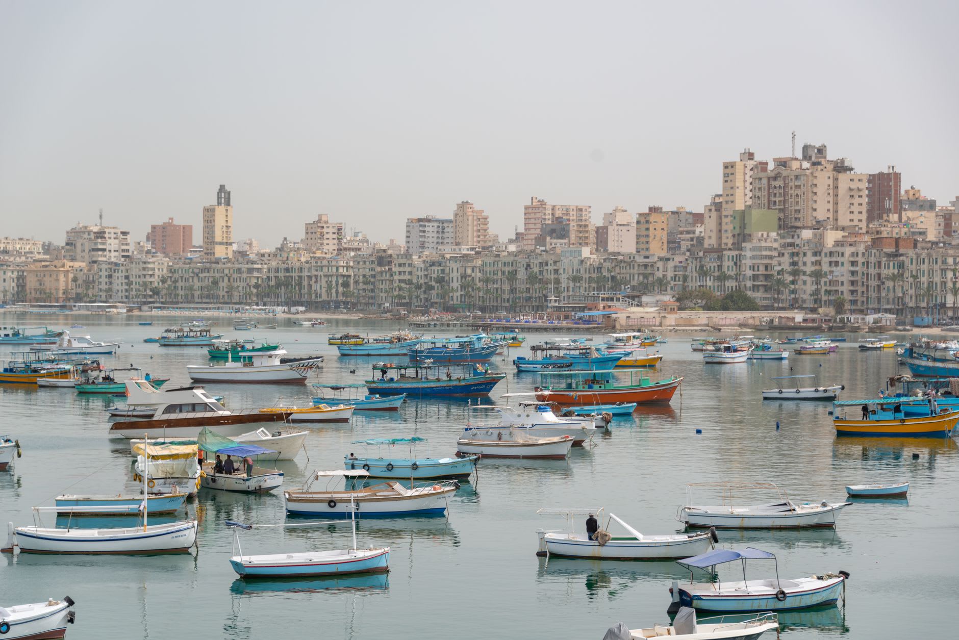 The corniche of Alexandria at golden hour, waves crashing against the seawall below faded colonial buildings