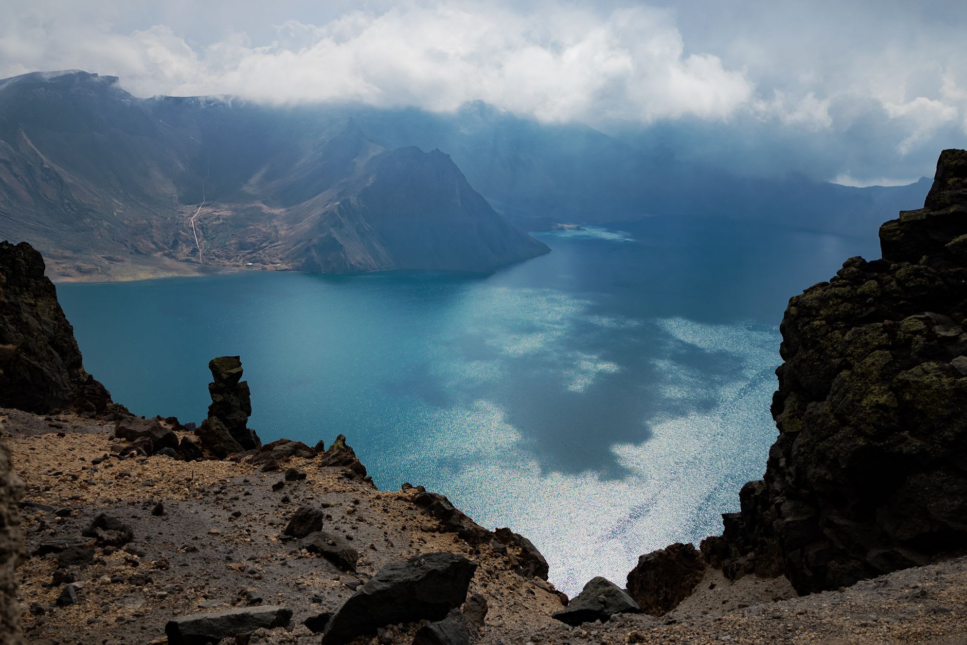 Lake Chon from the western rim, its crater walls reflecting in the deep blue water, the Chinese border peaks visible beyond