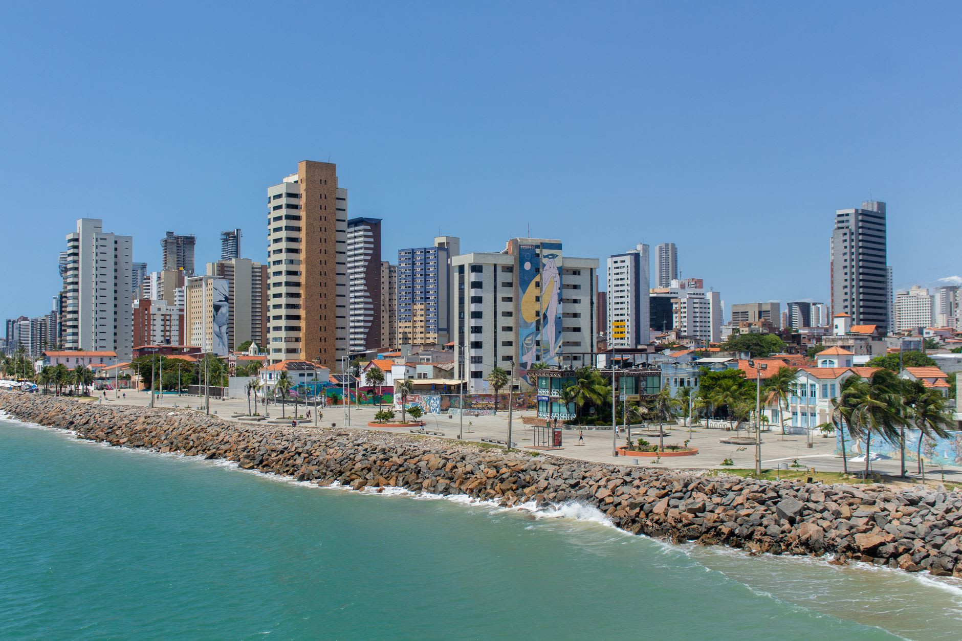 Dawn at Praia de Iracema in Fortaleza — fishermen at the rocky point, the sky warm orange and pink, a capoeira practitioner moving at the water's edge in the early light