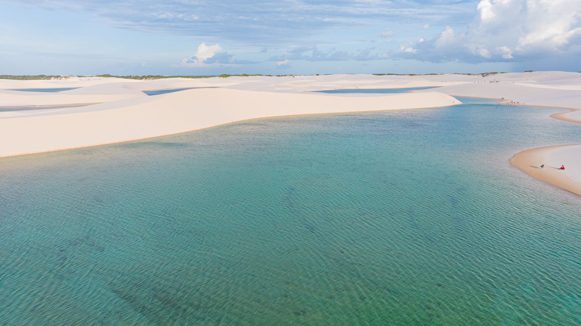A lone figure walking along a dune ridge at Lençóis Maranhenses at dusk, the orange sky reflected in the lagoon below