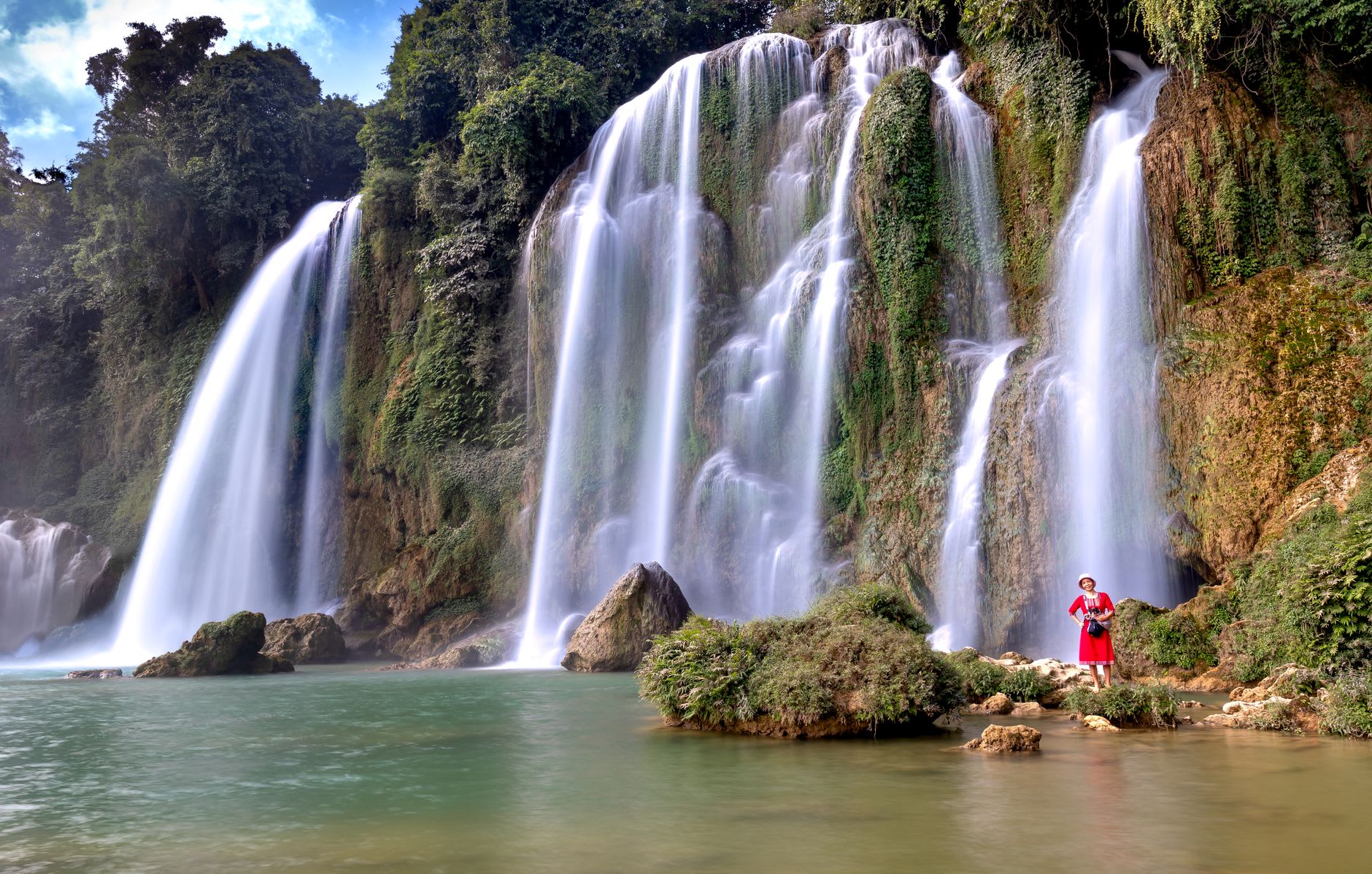 Bamboo raft on the turquoise pool at the base of Bản Giốc waterfall, the full width of the cascade filling the frame above, white water and green forest
