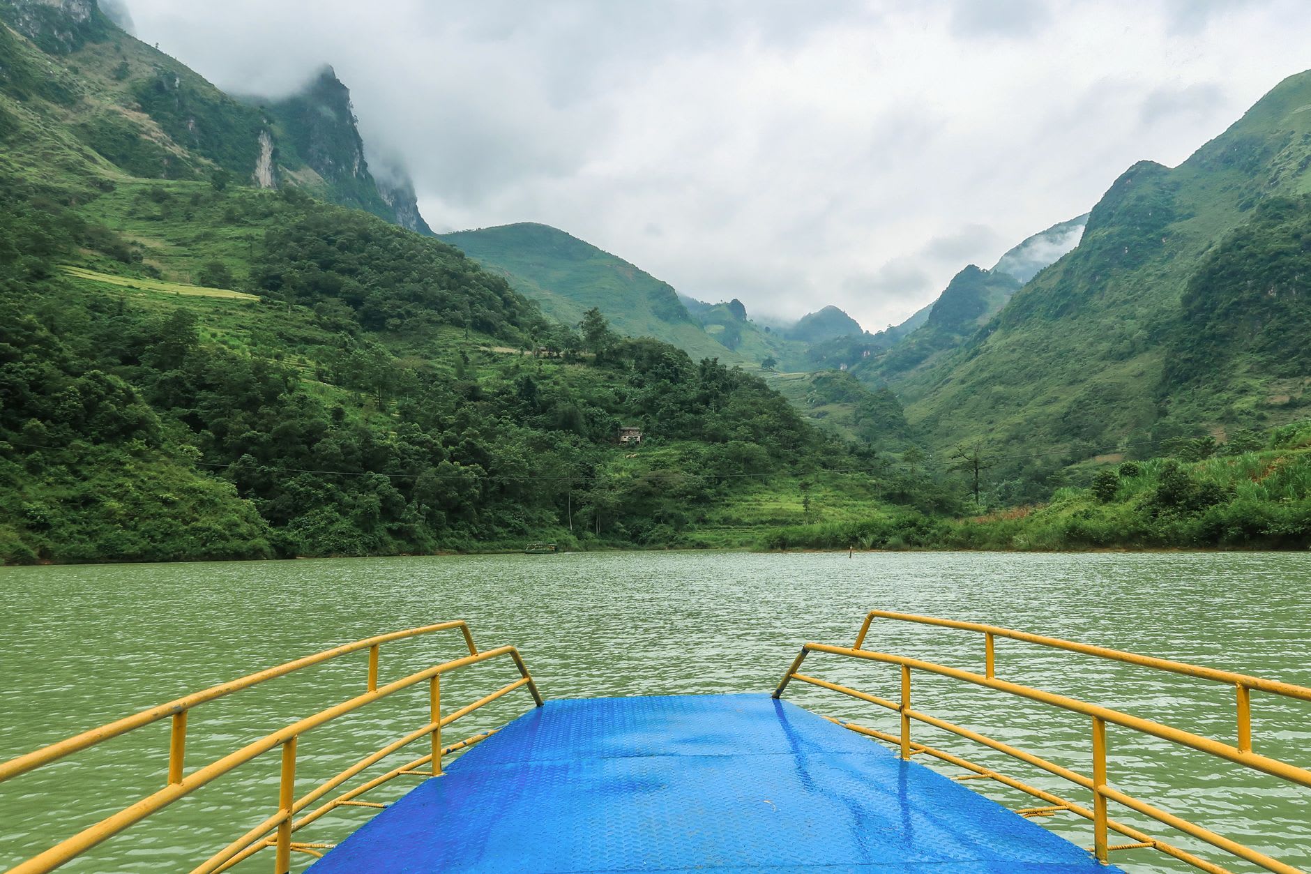 The Nho Que River at Meo Vac, deep jade green between limestone walls, a bamboo raft poled by a man in a conical hat in flat afternoon light