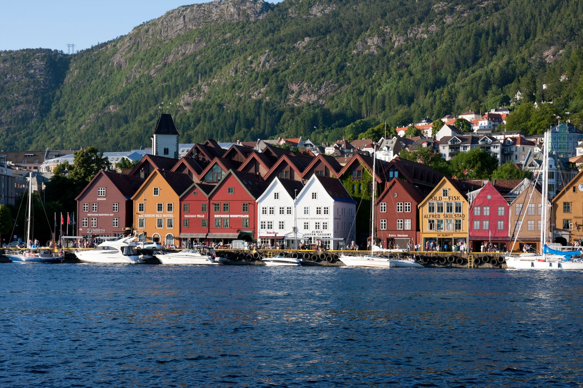 View from Mount Fløyen at dusk, Bergen city spread below in evening light, harbour and Bryggen waterfront visible, islands and archipelago extending into the distance