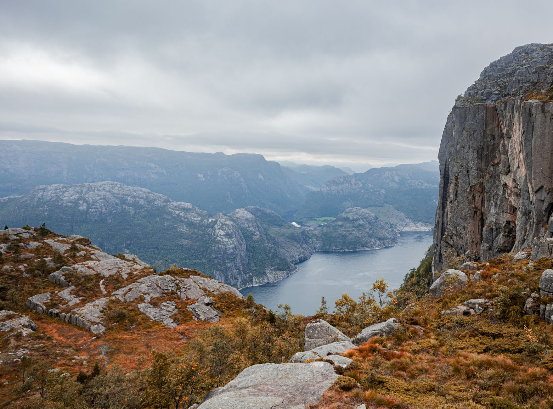 Looking down from the edge of Preikestolen, the vertical cliff face dropping 604 metres to the dark water of Lysefjord below, far wall of the fjord visible in the distance