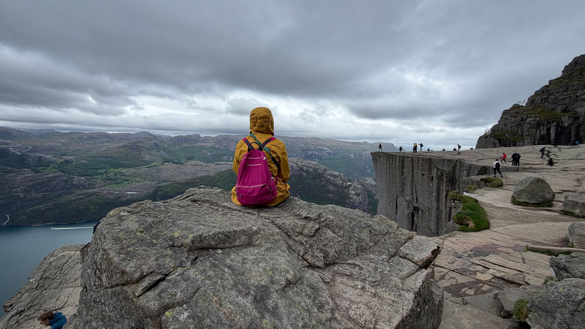 Wide view from Preikestolen at midday, hikers sitting along the edge, Lysefjord stretching toward the sea in the distance, scattered cloud shadows moving across the water below