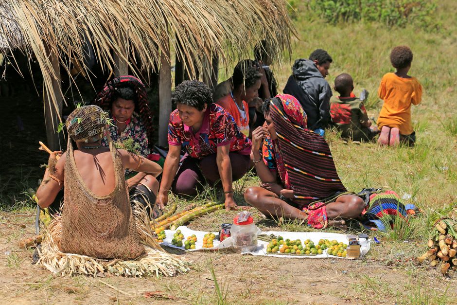 Le marché de produits d'Alotau en milieu de matinée, des vendeurs assis derrière des nattes tissées chargées de noix de bétel, de légumes verts et de fruits sous un toit en tôle ondulée