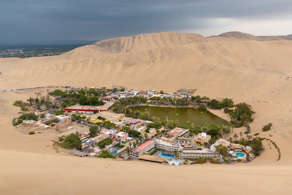 Sand dunes rising above a desert oasis with palm trees and still water