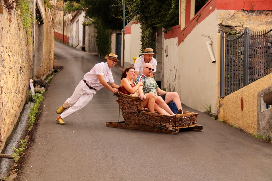 Two carreiro drivers in white and straw hats steering a wicker toboggan down Monte's cobblestone street