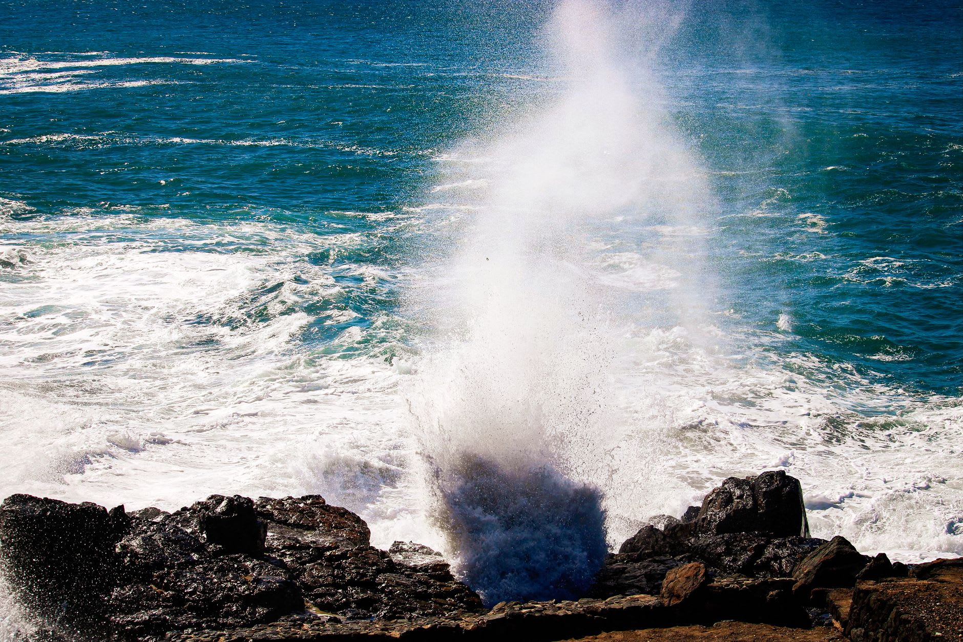 The main Alofaaga blowhole in full eruption, a column of white water against dark lava and the Pacific sky