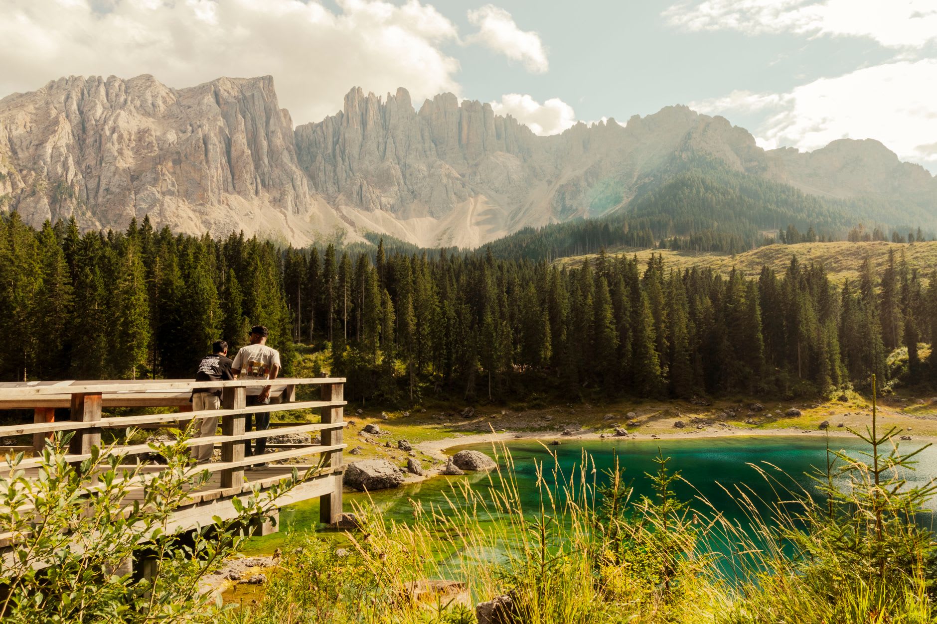 The pale jagged peaks of the Latemar range rising behind Lago di Carezza under shifting cloud light, dark forest framing the green water below