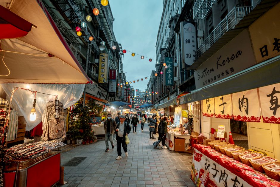 Vendors and lantern light along Danshui Old Street at dusk, the riverfront promenade filling with evening strollers