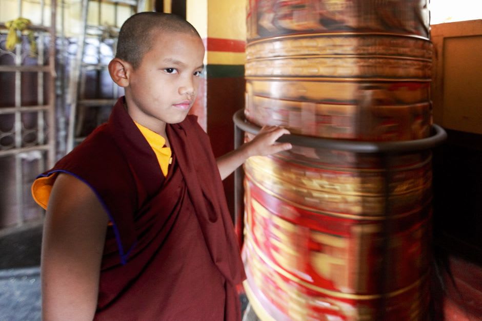 Maroon-robed monk crossing a stone courtyard at Ganden Monastery with prayer flags overhead