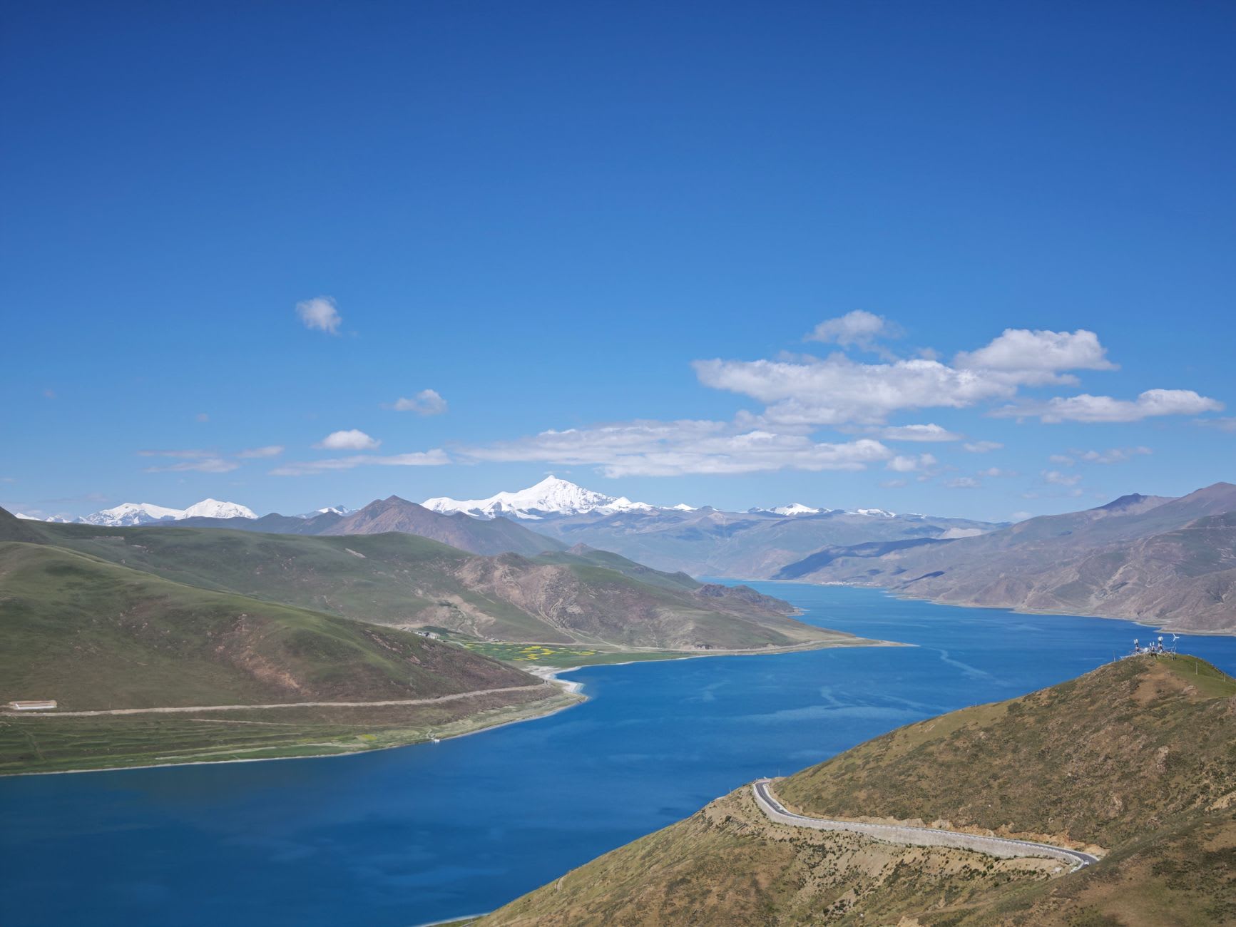 A stony shoreline at the edge of Yamdrok Lake with clear turquoise water shading from pale green shallows to deep blue, brown mountains beyond