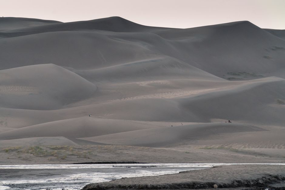 A hiker on a sharp dune ridge with wind lifting sand, mountains behind