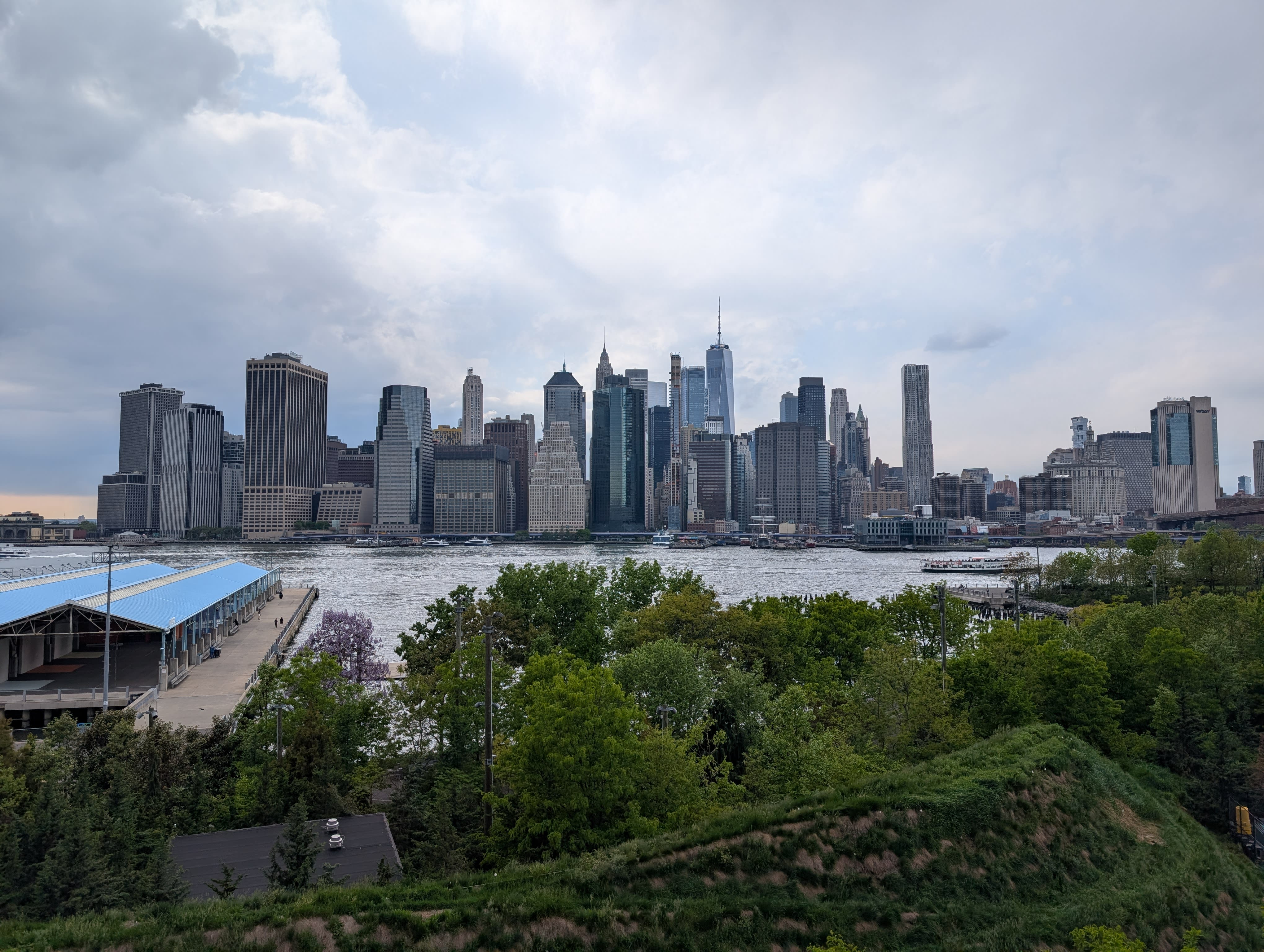 The Lower Manhattan skyline from Brooklyn Bridge Park — One World Trade Centre and the Financial District towers rising above the East River, green trees and park landscaping in the foreground, dramatic storm clouds gathering