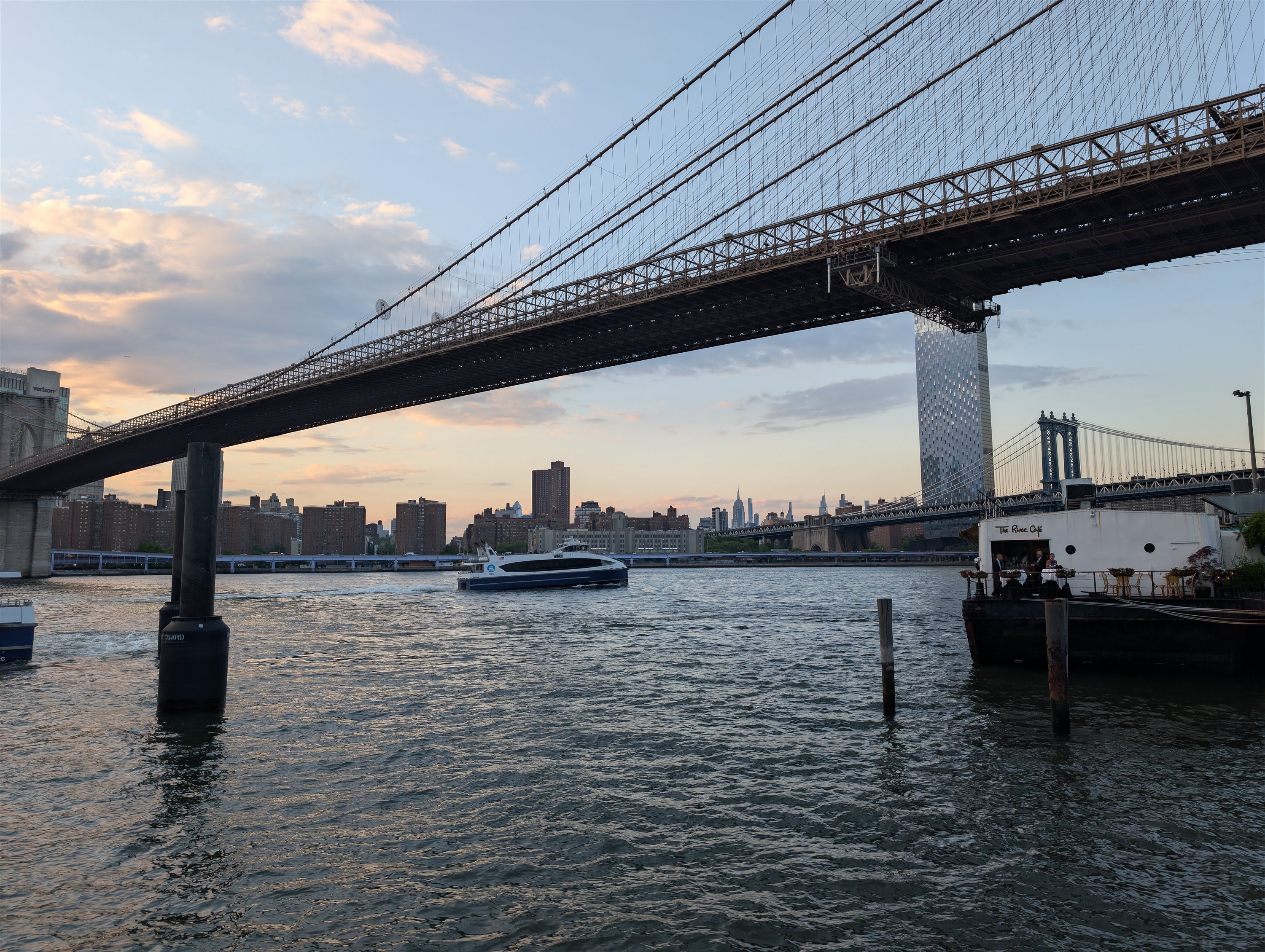 The Brooklyn Bridge at sunset — the steel structure silhouetted against a pink and gold sky, the Manhattan Bridge visible behind it, the East River reflecting the light, a ferry boat crossing the frame, the Empire State Building a thin spike on the distant horizon