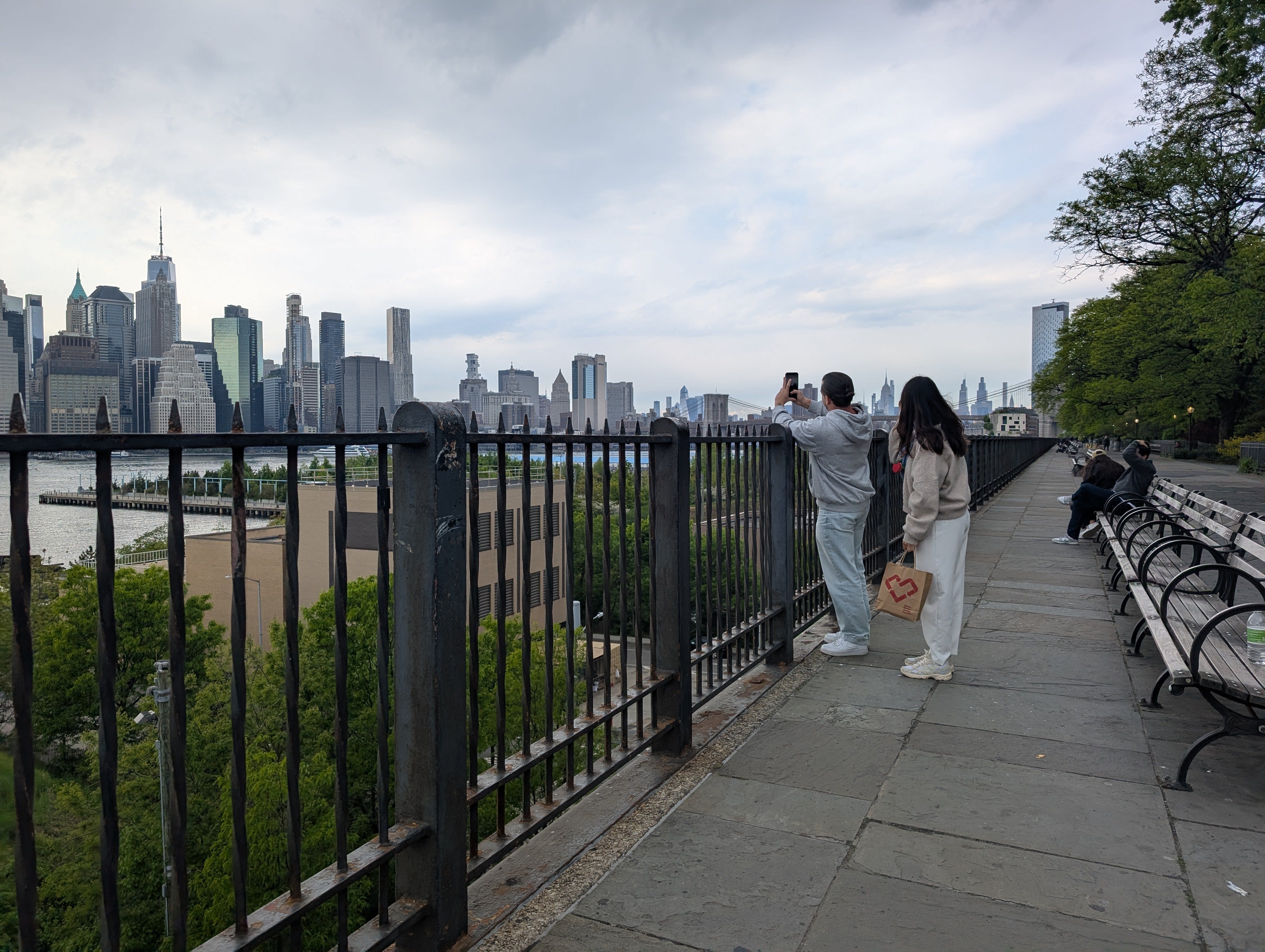 Pierre and Lia at the Brooklyn Heights Promenade railing, photographing the Manhattan skyline — the full Lower Manhattan cluster of towers across the East River, iron railing and wooden benches, overcast May sky