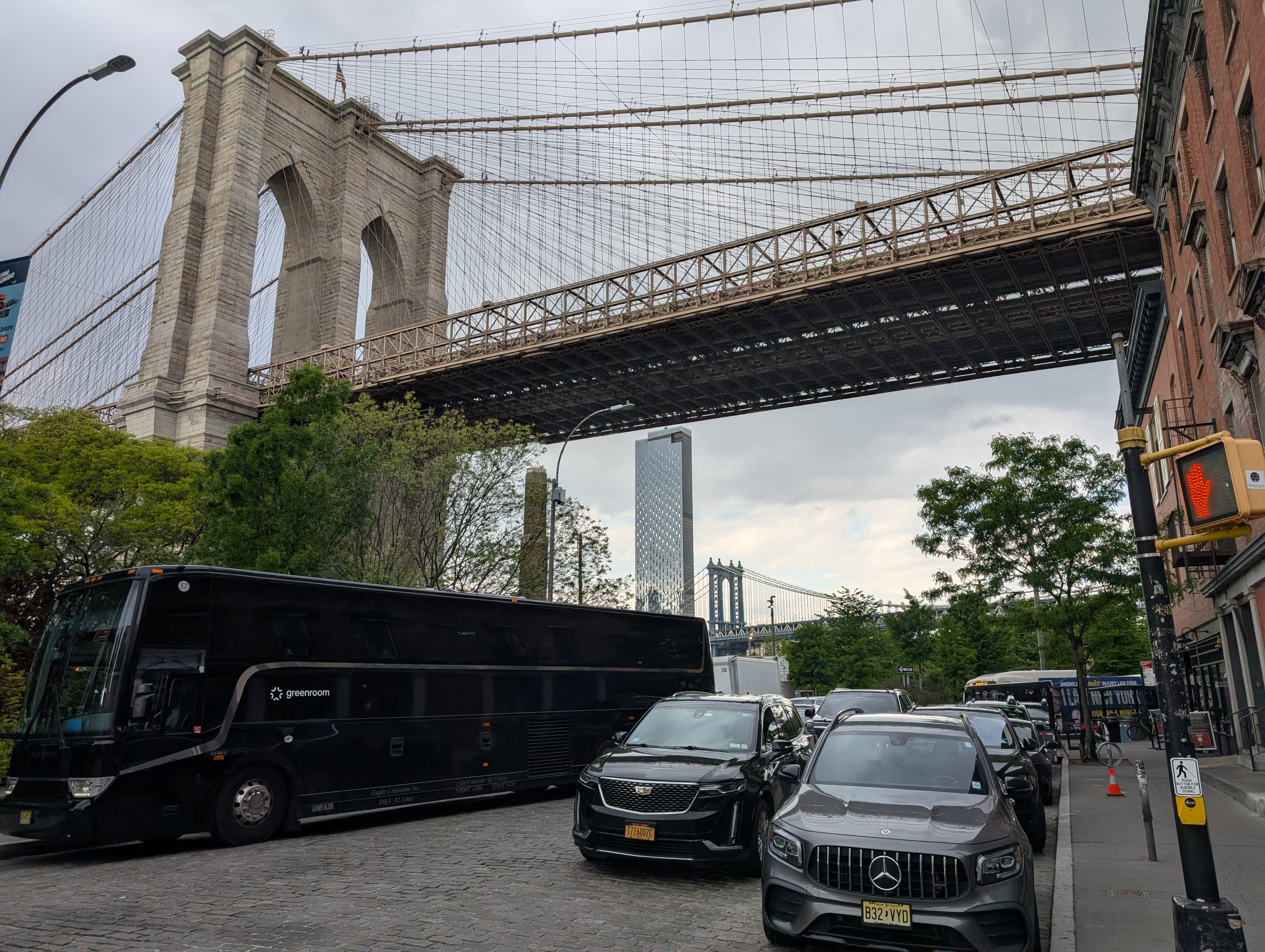 A DUMBO cobblestone street — the Brooklyn Bridge overhead, the Manhattan Bridge tower visible in the gap between buildings, a black tour bus and luxury vehicles parked on the stones, the compressed geometry of old warehouse Brooklyn
