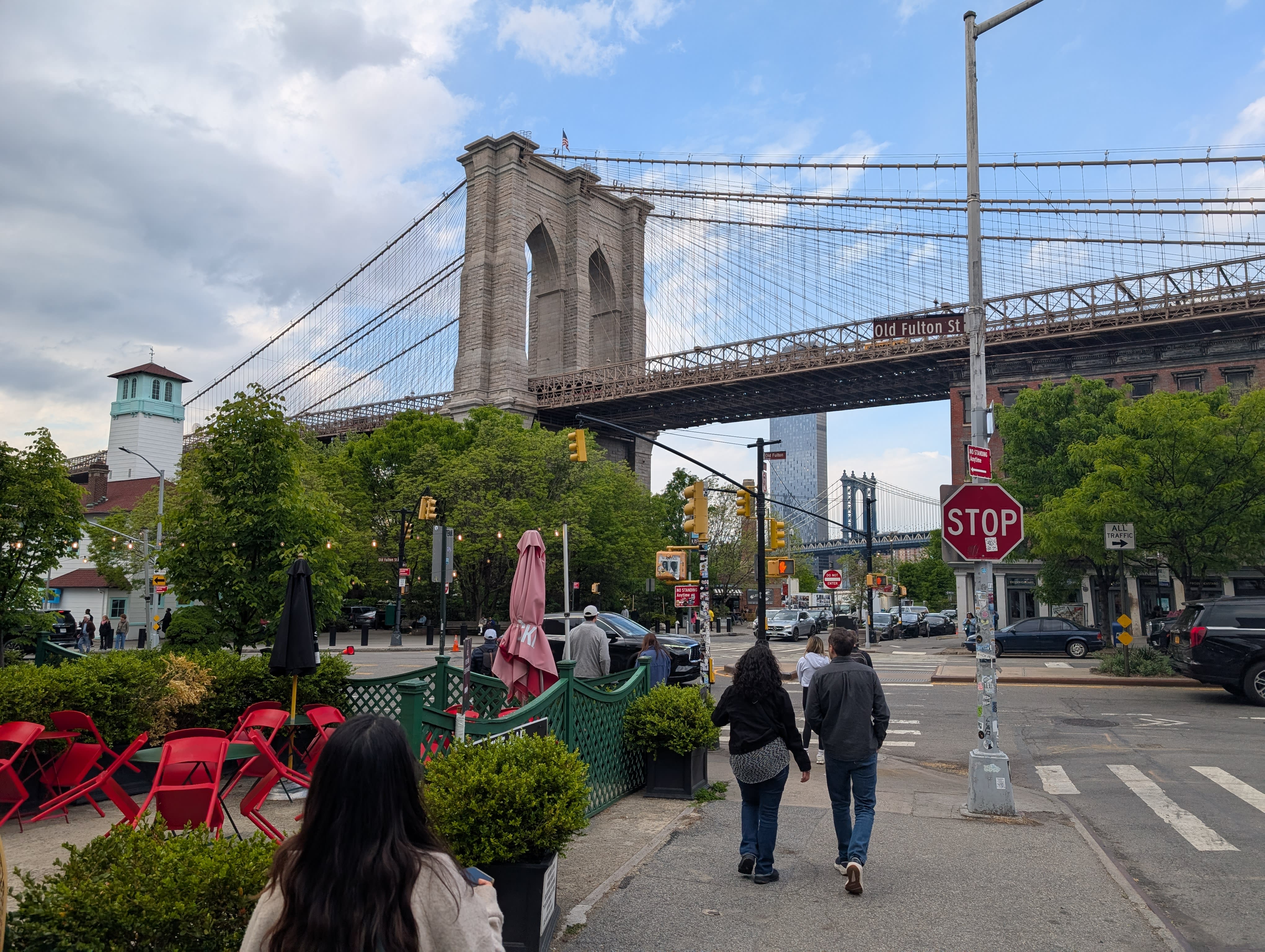 Old Fulton Street in DUMBO — the Brooklyn Bridge stone tower rising above the trees, the Manhattan Bridge visible in the distance behind it, red café chairs on the left, a stop sign, pedestrians walking toward the bridge