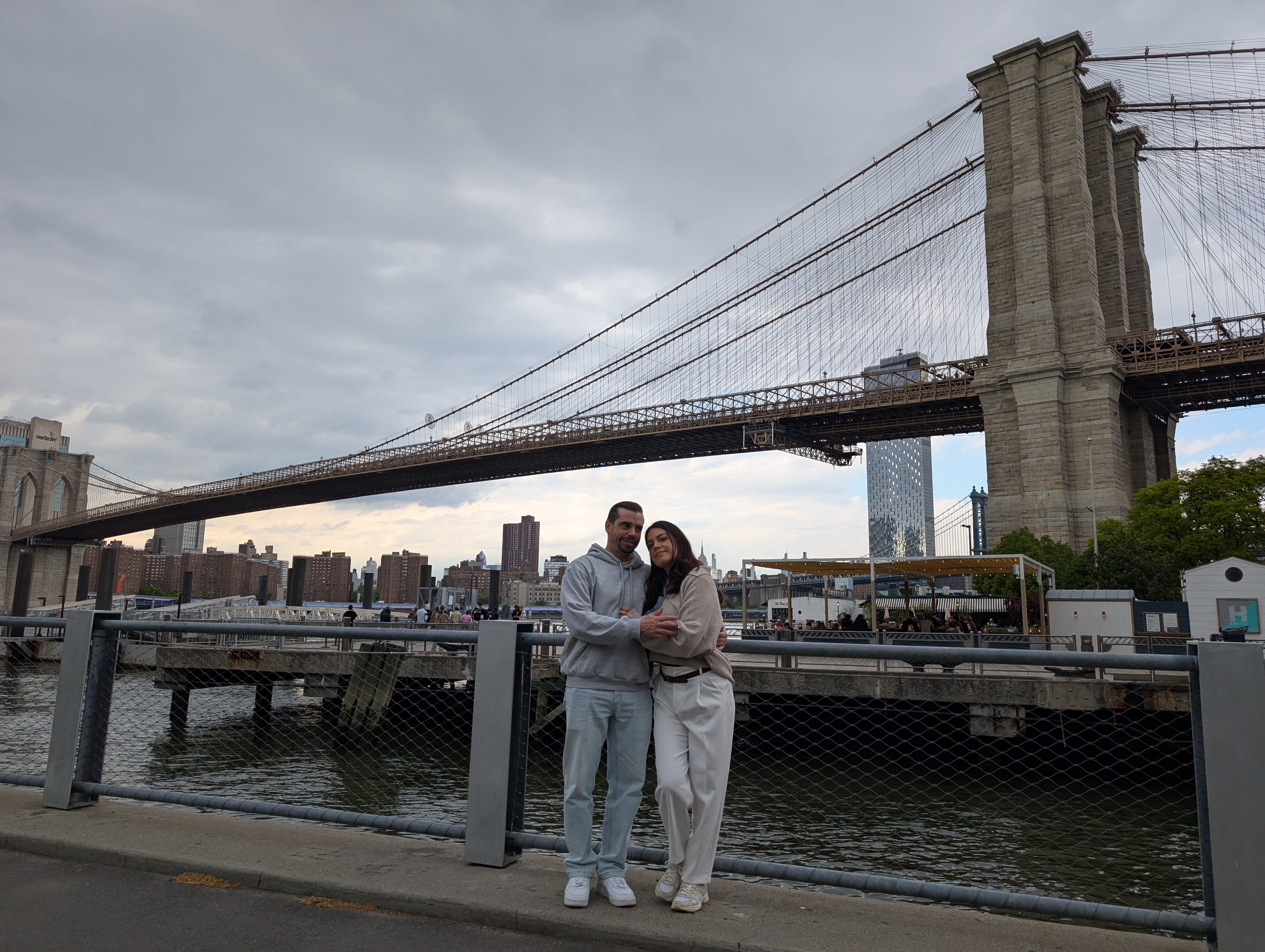 Pierre and Lia embracing at the DUMBO waterfront railing — the Brooklyn Bridge spanning overhead in its full length, the East River and Manhattan skyline behind them, overcast sky