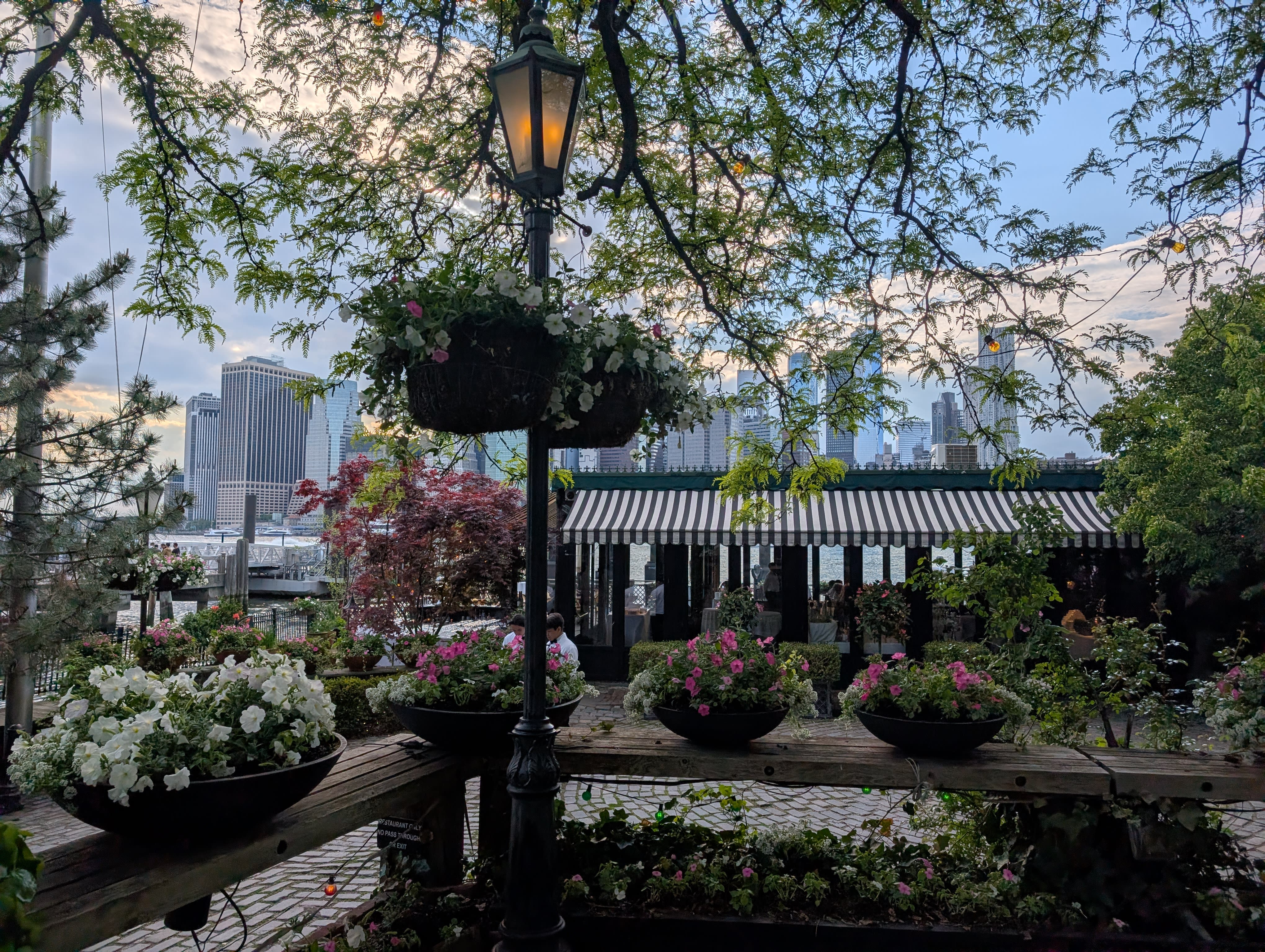 The River Café garden terrace at golden hour — hanging flower baskets, a wrought-iron gas lamp glowing, potted flowers in iron bowls, a black-and-white striped awning, and through the trees the Lower Manhattan skyline catching the evening light