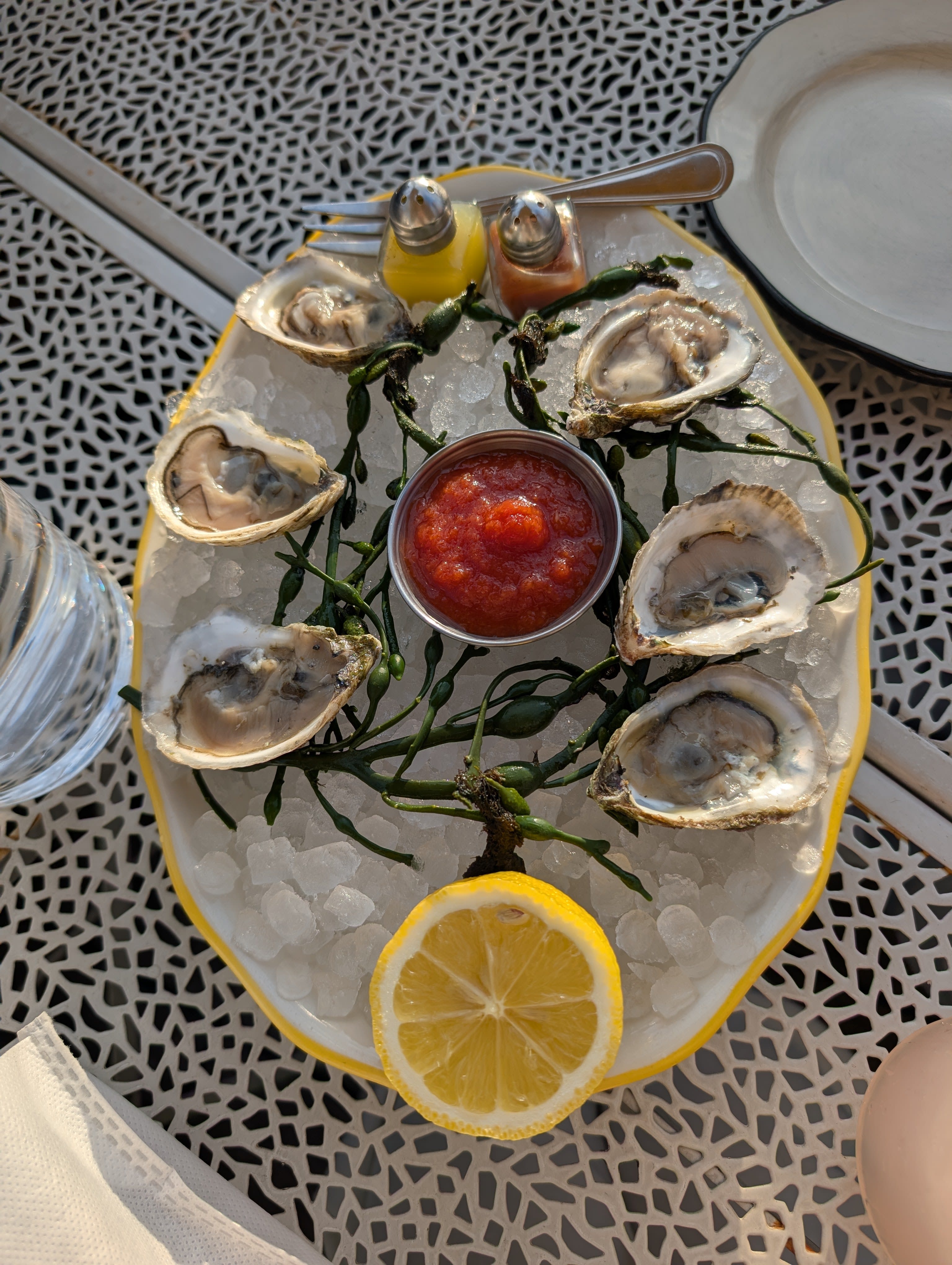 Six fresh oysters on a bed of crushed ice — arranged on a yellow-rimmed platter with samphire, a ramekin of cocktail sauce, a halved lemon, and the small fork, on a patterned metal table