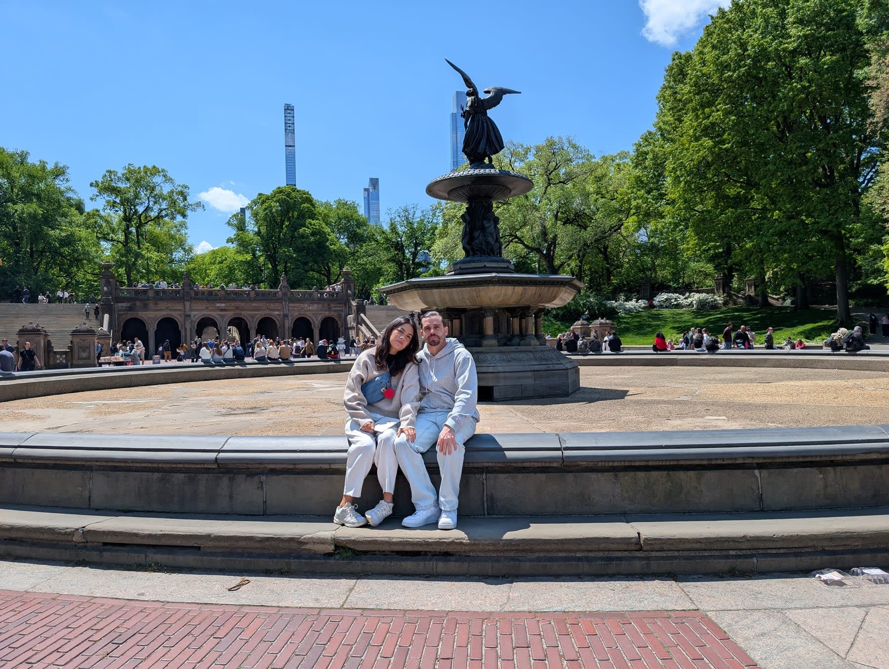 Lia and Pierre close together at the fountain's edge, the Angel of the Waters centred directly behind them, the sandstone terrace and the supertalls of the 57th Street skyline visible on the horizon above the green canopy — a clear May afternoon at the heart of the park