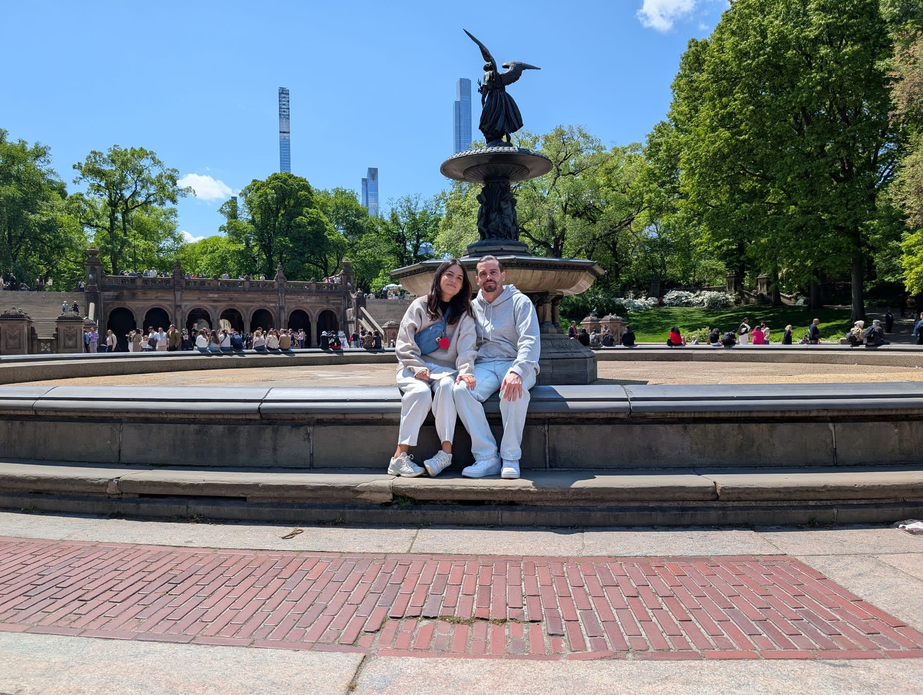 Lia and Pierre sitting together on the edge of Bethesda Fountain — the Angel of the Waters rising behind them, the sandstone arcade of the terrace visible in the background, the park's May-green trees on both sides, clear blue sky above, the supertalls of Billionaires' Row thin on the horizon