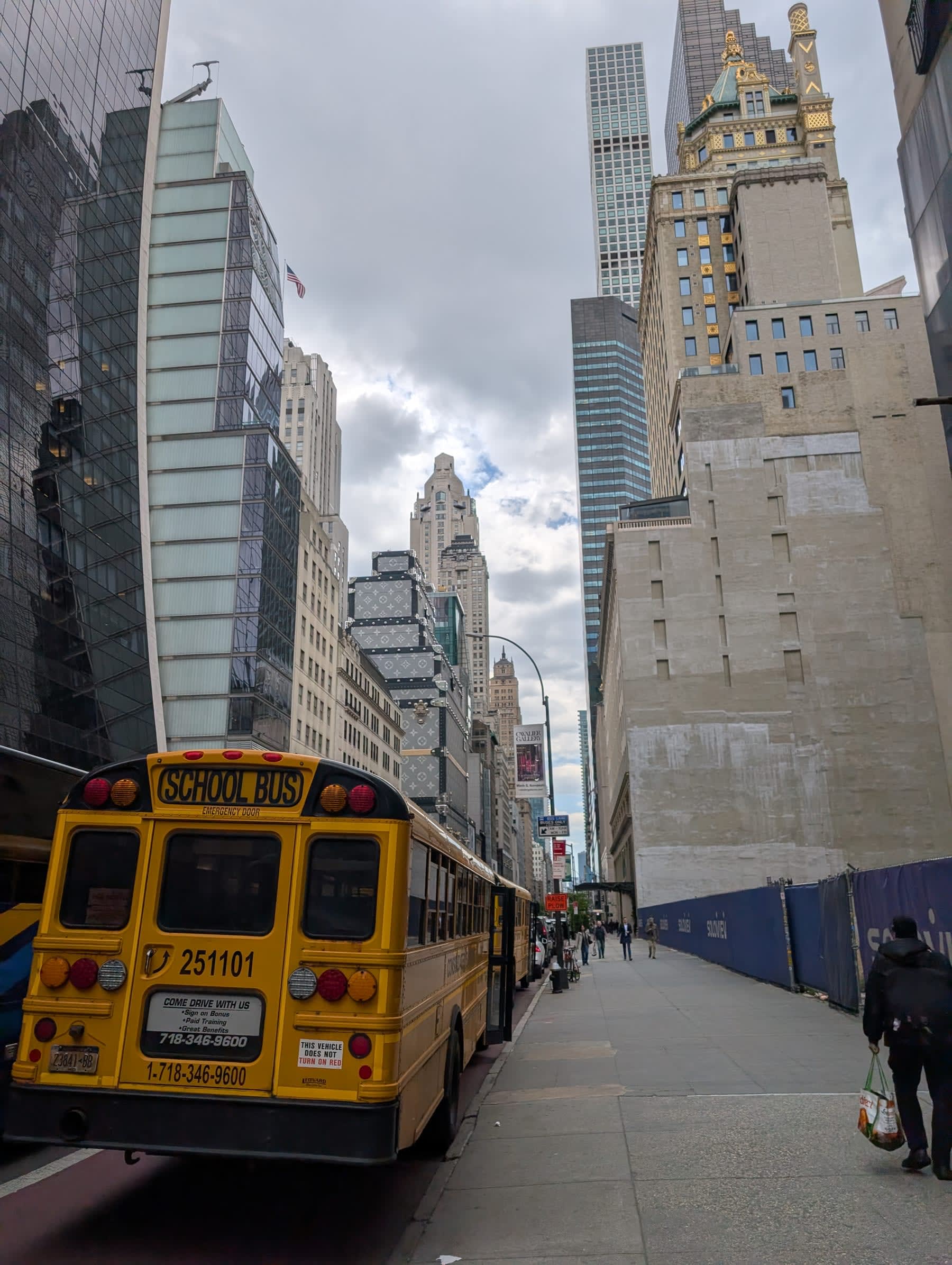 Fifth Avenue looking north toward 59th Street — a yellow school bus in the right lane, midtown towers rising on both sides, the Sherry-Netherland's gilded roof visible above the canyon, a grey May sky overhead